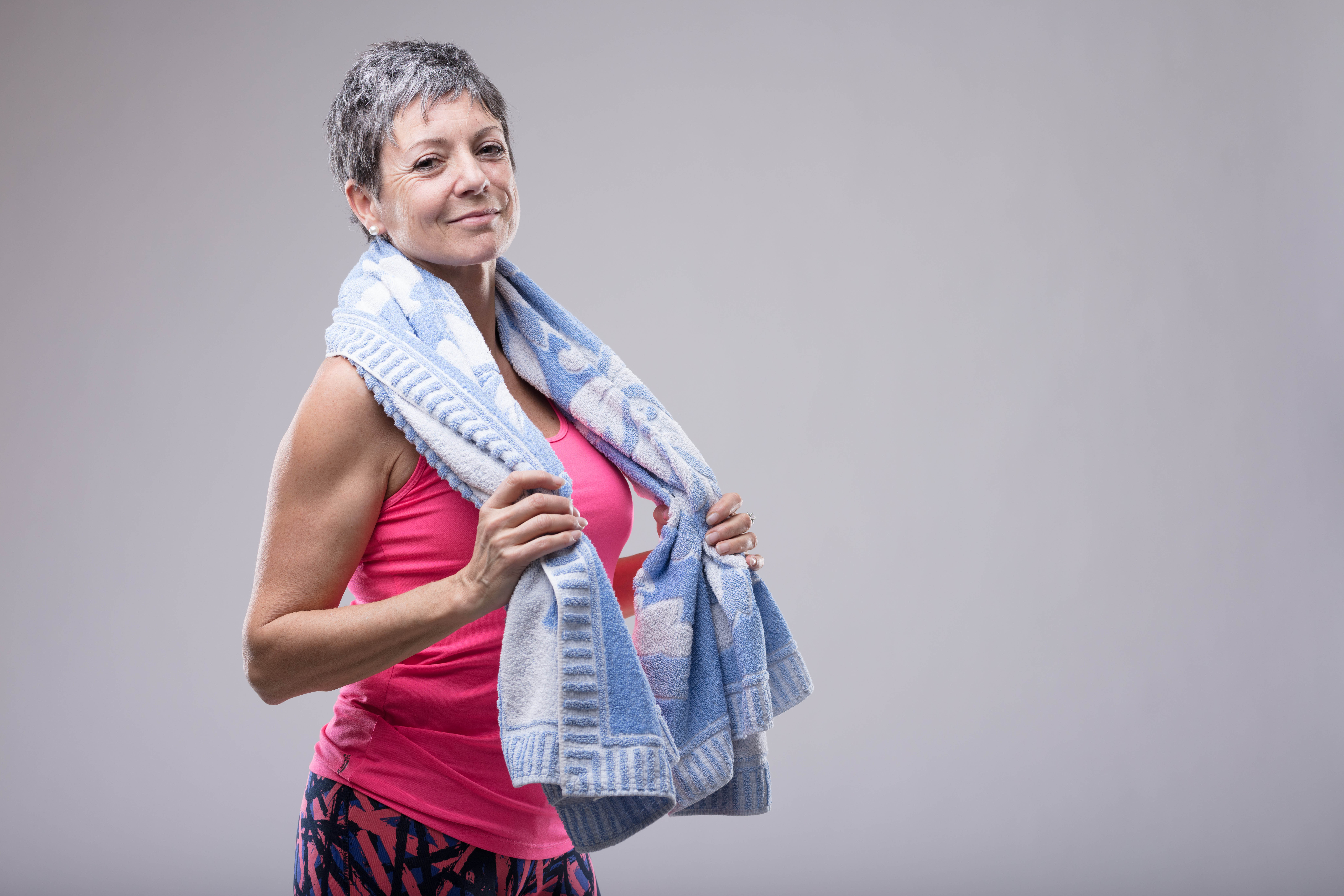 Older woman dressed in pink workout gear, standing with a towel wrapped around her neck, hands holding the towel, preparing for a workout